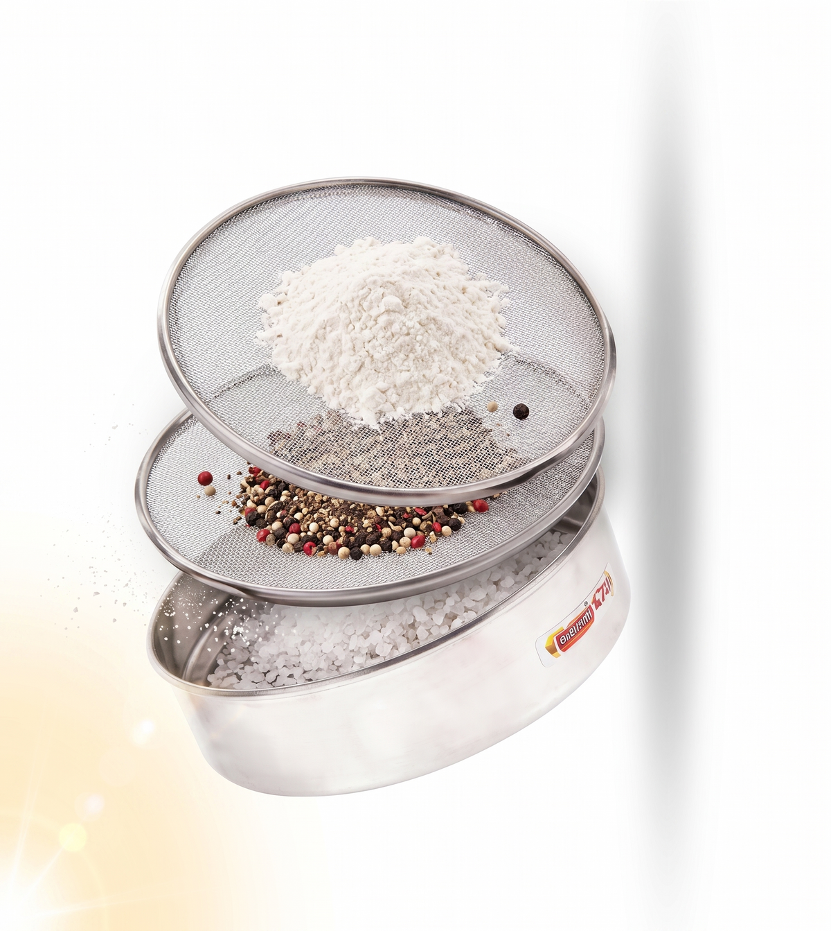 Three overlapping stainless steel sifter sieves with mesh screens, shown floating on a white background. The sieves contain white flour, coarse salt, and a mixed peppercorn pile.